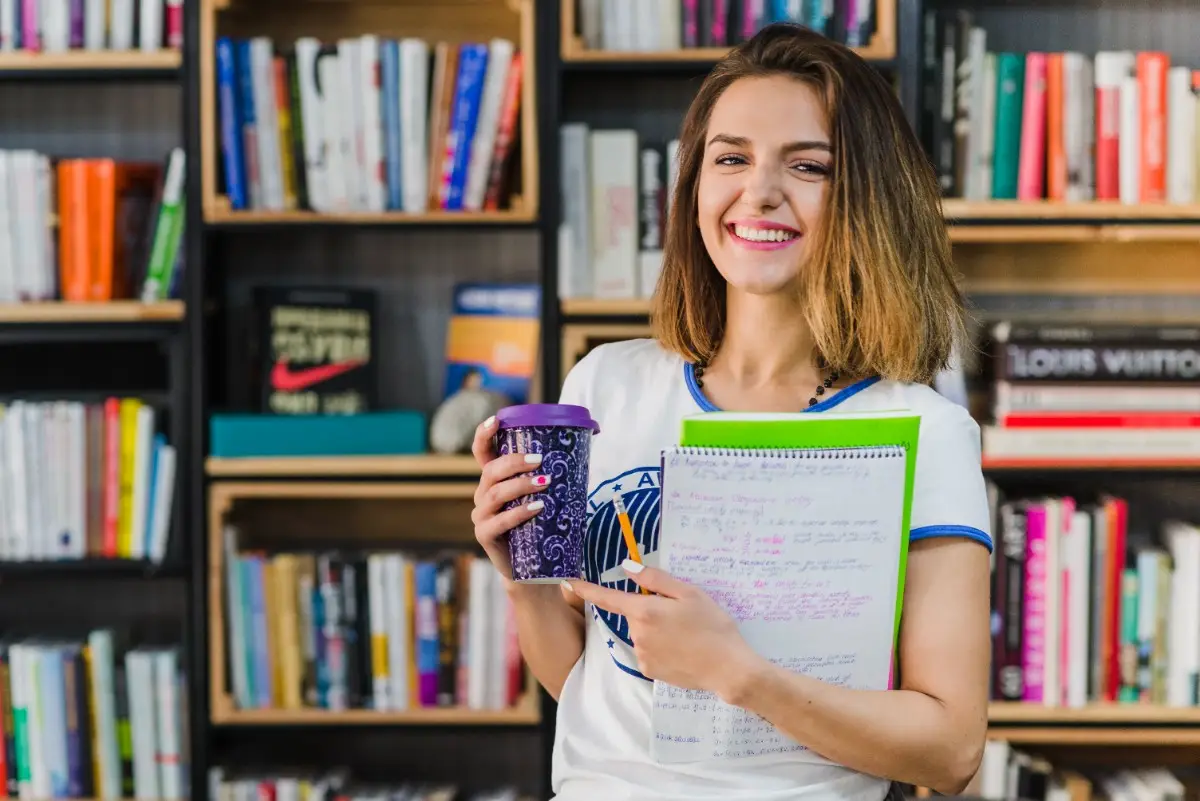 Smiling young woman holding a notebook and coffee cup at a library or study centre, searching for a german coaching centre near me
