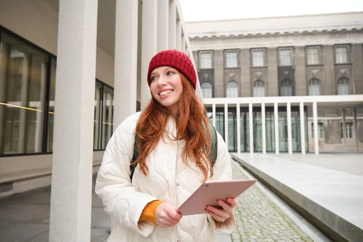 Best German language education consultancy in Kerala: Smiling young woman with red hair and knit cap holding a tablet on a university campus
