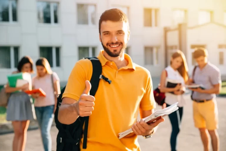 Smiling young man with a backpack holding books and giving a thumbs-up in front of a campus, representing students at an affordable German language institute in Kochi, Kerala.