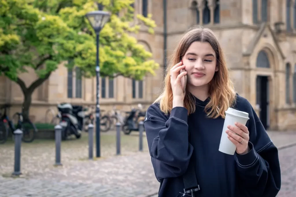 Student exploring Ausbildung in Germany for Indians from Kerala while walking near a German university campus with a coffee and phone.